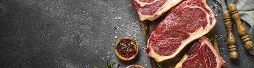 Raw meat beef steak. Black angus prime meat set - ribeye, striploin, t-bone steaks on cutting board. Top view on black table with copy space.